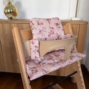 May include: A wooden high chair featuring a light pink fabric cushion set with a floral pattern of red roses and green leaves. The chair includes a beige plastic safety rail and backrest support, with a natural wood frame and adjustable seat slots.