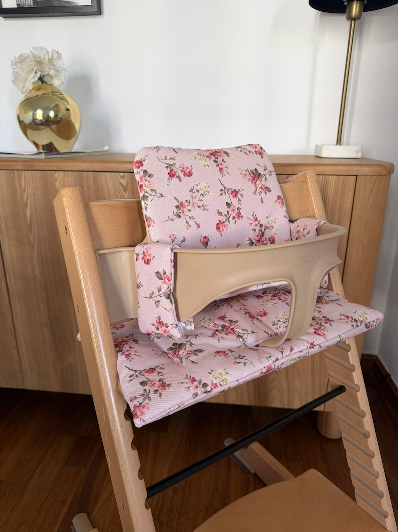 May include: A wooden high chair featuring a light pink fabric cushion set with a floral pattern of red roses and green leaves. The chair includes a beige plastic safety rail and backrest support, with a natural wood frame and adjustable seat slots.
