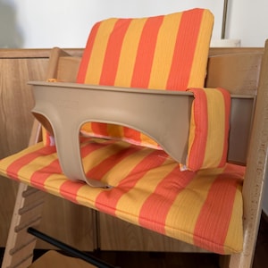 May include: A wooden high chair with a beige plastic safety bar and orange and yellow striped cushions. The chair has a footrest and is designed for a child. The cushions are padded and provide comfort.
