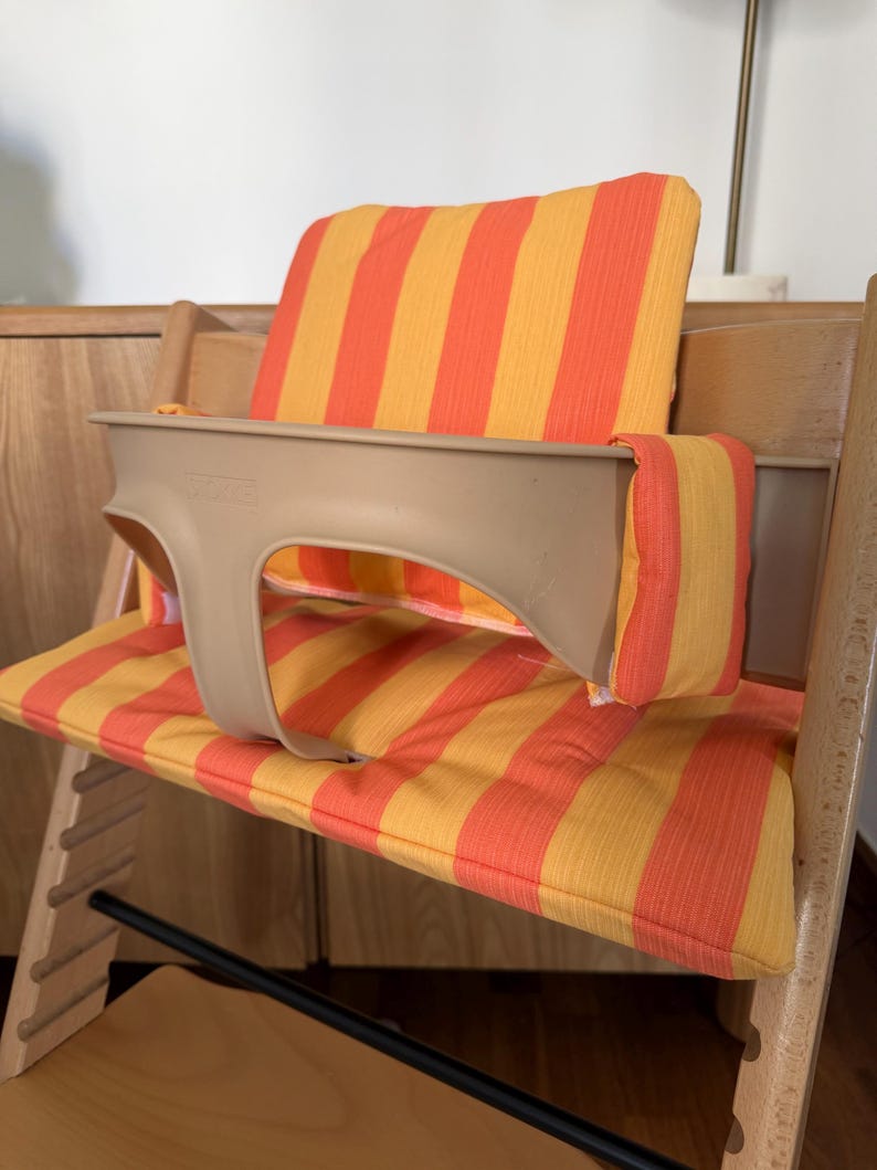 May include: A wooden high chair with a beige plastic safety bar and orange and yellow striped cushions. The chair has a footrest and is designed for a child. The cushions are padded and provide comfort.