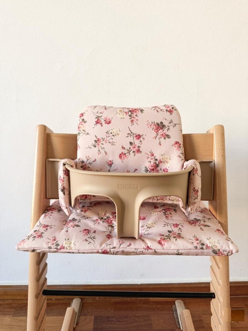 May include: A light-colored wooden high chair featuring a beige plastic safety rail with the embossed text STOKKE. The chair includes a soft, padded seat and backrest cushion covered in a light pink fabric with a floral pattern of red roses.