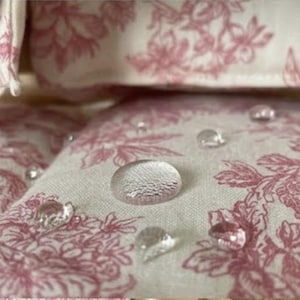 May include: A light-colored fabric featuring a repeating pink floral pattern with several clear water droplets resting on the surface, demonstrating a water-repellent quality of the textile material with visible woven texture.