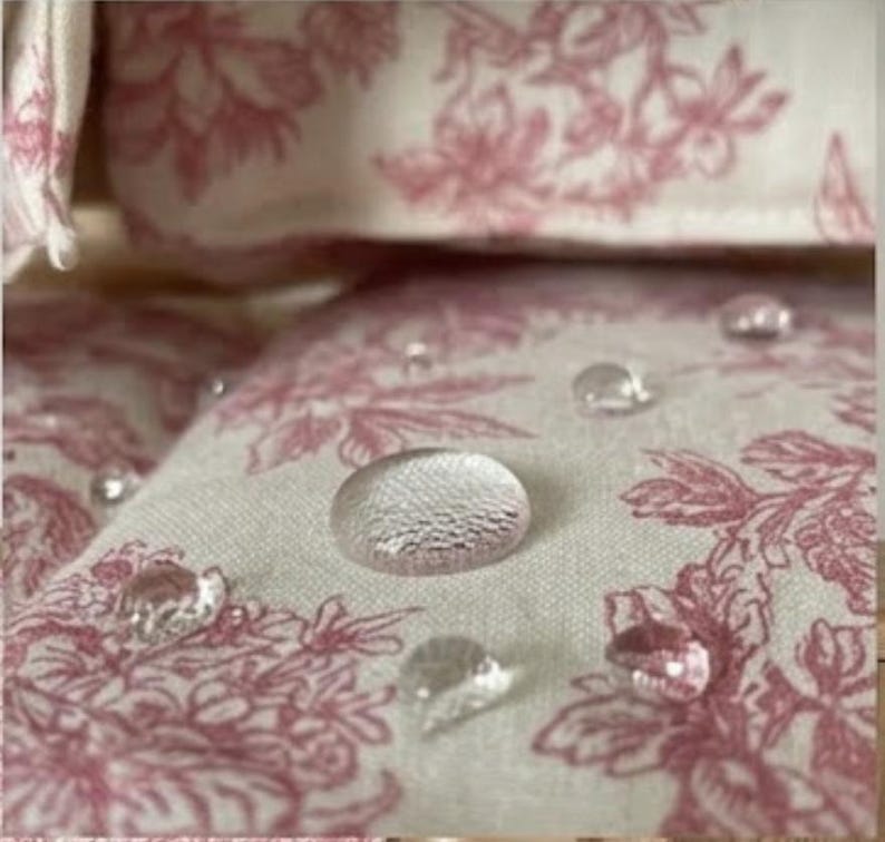 May include: A light-colored fabric featuring a repeating pink floral pattern with several clear water droplets resting on the surface, demonstrating a water-repellent quality of the textile material with visible woven texture.
