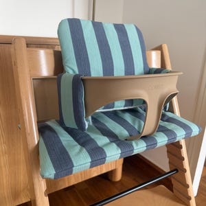 May include: A wooden high chair featuring a light beige plastic safety rail with the word STOKKE embossed on the front. The chair includes a padded seat and backrest cushion with a vertical striped pattern in alternating shades of teal and dark grey.