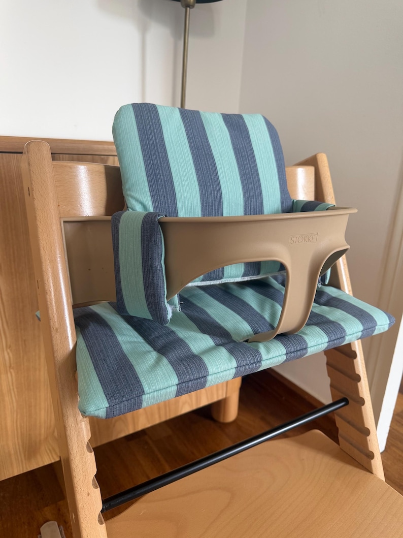 May include: A wooden high chair featuring a light beige plastic safety rail with the word STOKKE embossed on the front. The chair includes a padded seat and backrest cushion with a vertical striped pattern in alternating shades of teal and dark grey.