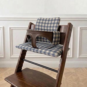 May include: A dark brown wooden high chair featuring a blue and cream plaid fabric cushion set. The chair includes a dark brown plastic safety guard and a footrest. The seat and backrest are padded with the plaid fabric, providing support.
