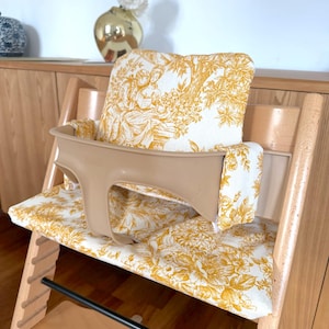 May include: A wooden high chair featuring a beige plastic safety rail and a padded seat cushion set with a white and mustard yellow toile de jouy pattern. The fabric displays intricate illustrations of figures and foliage in a repeating design.