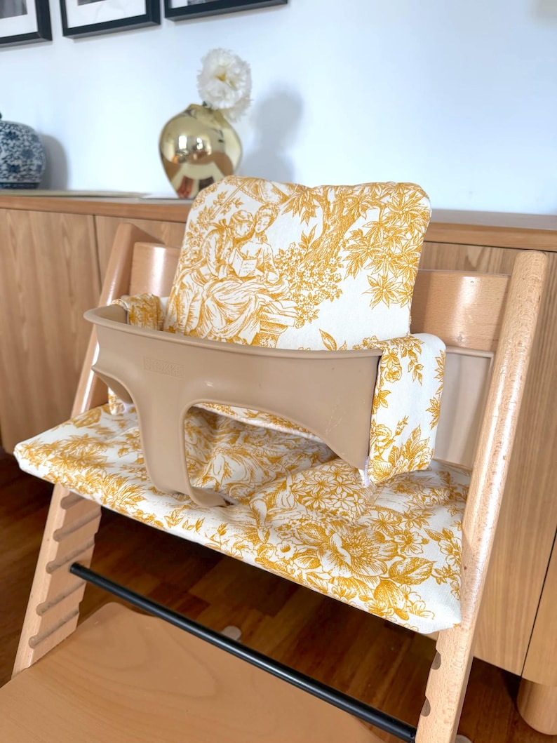 May include: A wooden high chair featuring a beige plastic safety rail and a padded seat cushion set with a white and mustard yellow toile de jouy pattern. The fabric displays intricate illustrations of figures and foliage in a repeating design.