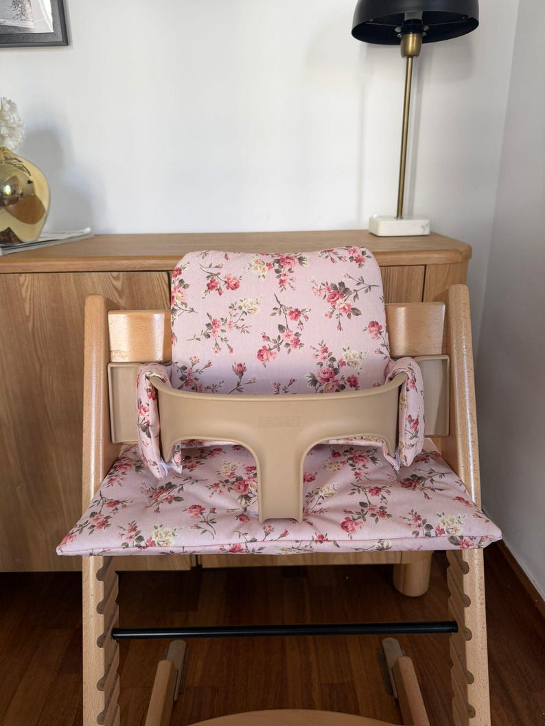 May include: A wooden high chair featuring a light pink fabric cushion set with a floral pattern of red roses and green leaves. The chair includes a beige plastic safety guard and backrest support, with a natural wood frame and a black footrest bar.