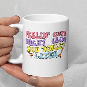 May include: White ceramic mug with a pink, yellow, and blue graphic that reads "Feelin' Cute Might Clog The Toilet Later" with two toilet plunger icons.