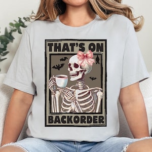 Könnte beinhalten: Ein graues T-Shirt mit einem Motiv eines Skeletts, das eine Kaffeetasse hält und eine rosa Schleife trägt. Der Text "That's on Backorder" ist unter dem Skelett gedruckt.