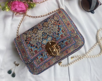 May include: A small, patterned handbag with a gold chain strap and a gold clasp. The bag has a colorful, intricate design with a blue, red, and gold color scheme.
