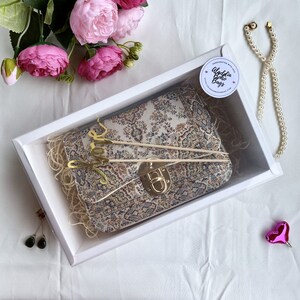 May include: A patterned clutch bag with a gold clasp, presented in a white box with decorative straw. The bag has a beige, brown, and green floral design. The box is decorated with pink flowers, a pearl necklace, and a pair of earrings.