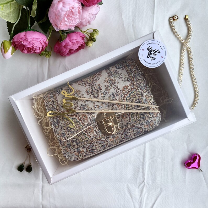 May include: A patterned clutch bag with a gold clasp, presented in a white box with decorative straw. The bag has a beige, brown, and green floral design. The box is decorated with pink flowers, a pearl necklace, and a pair of earrings.