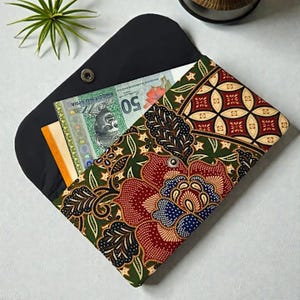 May include: A black clutch wallet with a colorful floral batik pattern in red, green, and blue. The wallet is open, revealing Malaysian currency and a small stack of cards. The wallet is closed with a snap closure.