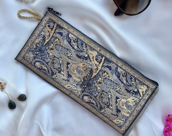 May include: A rectangular, navy blue paisley pattern glasses case with a gold zipper and a gold cord loop. The case features a detailed floral design in gold and light blue. Sunglasses are visible in the background.