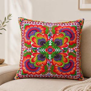 Puede incluir: Un cojín cuadrado con bordados simétricos y vibrantes en tonos naranja, rojo, morado y verde. El diseño presenta motivos florales y de hojas intrincados dispuestos en un patrón central, rodeado por un borde geométrico decorativo.