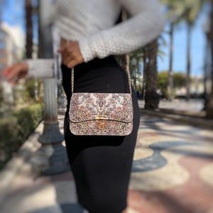 May include: A rectangular handbag with a decorative floral pattern in beige, brown, and orange hues. The bag features a gold-colored clasp and chain strap. The bag is approximately 9 inches wide and 6 inches tall.