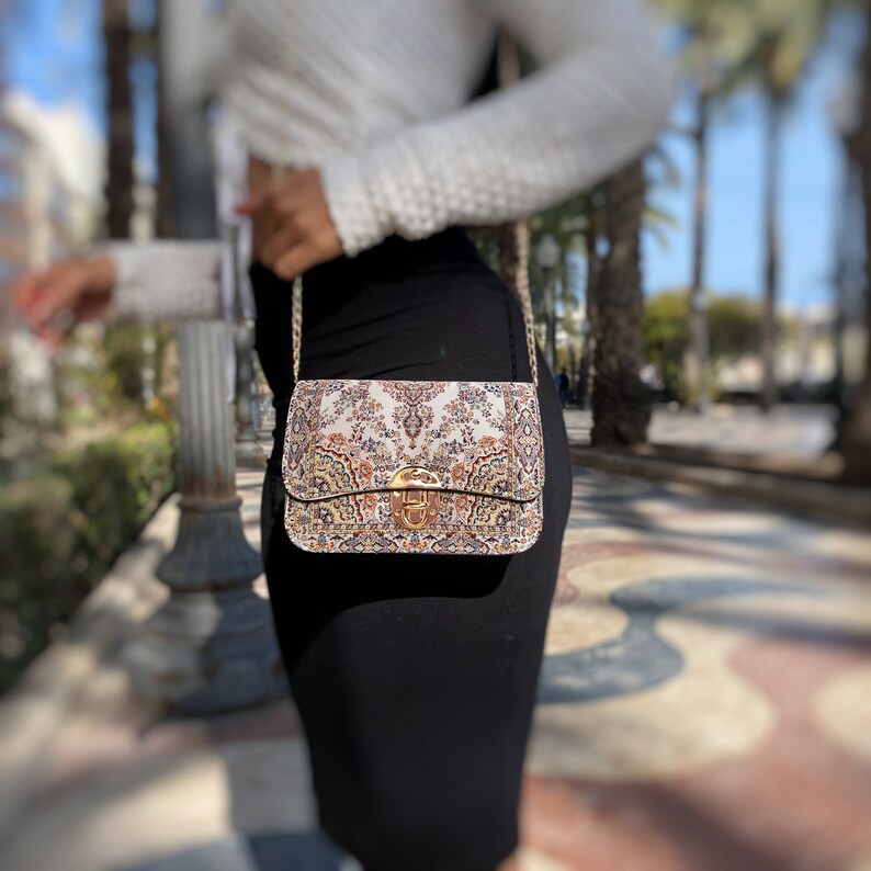 May include: A rectangular handbag with a decorative floral pattern in beige, brown, and orange hues. The bag features a gold-colored clasp and chain strap. The bag is approximately 9 inches wide and 6 inches tall.