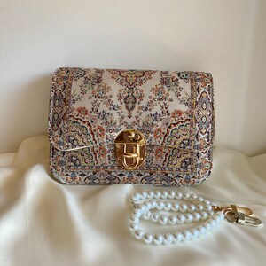 May include: A rectangular handbag with a tapestry-like pattern in cream, red, blue, and gold. The bag features a gold-colored clasp and a pearl beaded wrist strap with a gold-colored clasp.