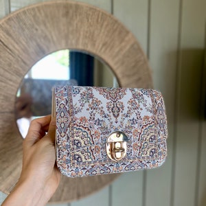May include: A rectangular handbag with a patterned fabric design in shades of cream, blue, and rust. The bag features a gold-colored clasp and a structured shape. The bag is held up in front of a round mirror.