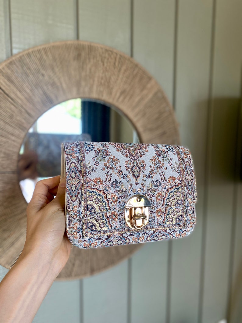 May include: A rectangular handbag with a patterned fabric design in shades of cream, blue, and rust. The bag features a gold-colored clasp and a structured shape. The bag is held up in front of a round mirror.