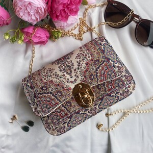 May include: A rectangular handbag with a gold chain strap and clasp. The bag features a cream-colored background with a detailed pattern in red, blue, and gold. Sunglasses and jewelry are also visible.