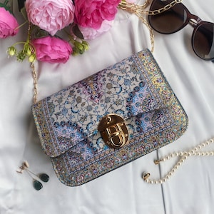 May include: A rectangular handbag with a gold chain strap and a gold clasp. The bag features a floral pattern in shades of blue, pink, and gold. The background includes pink flowers, sunglasses, earrings, and a pearl necklace.