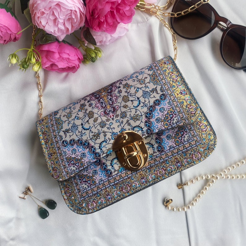May include: A rectangular handbag with a gold chain strap and a gold clasp. The bag features a floral pattern in shades of blue, pink, and gold. The background includes pink flowers, sunglasses, earrings, and a pearl necklace.