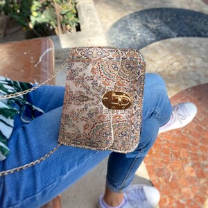 May include: A rectangular handbag with a gold chain strap and a gold clasp. The bag features a beige, brown, and orange floral pattern. The bag is resting on a person's lap, with blue jeans and white sneakers visible.