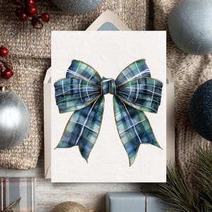 Pode incluir: Um cartão com uma ilustração em aquarela de um laço xadrez azul e verde com acabamento dourado. O cartão está sobre um fundo neutro com enfeites de Natal e bagas.