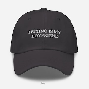 May include: A grey baseball cap featuring the white embroidered text "TECHNO IS MY BOYFRIEND". The cap has a curved peak and an adjustable strap. The word "Grey" is printed at the bottom of the cap.