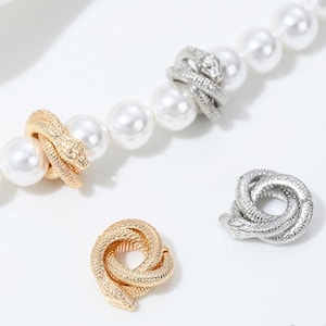 May include: Three gold and silver snake-shaped charms. The charms are in the shape of a snake coiled around itself. The charms are on a white pearl necklace.