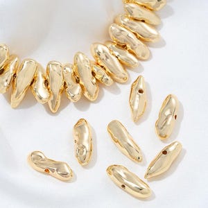 Può includere: Collana e perline sciolte color oro. La collana presenta perline allungate e irregolari. Diverse perline sciolte sono sparse intorno alla collana. Le perline hanno un aspetto strutturato e organico, che riflette la luce. Lo sfondo è una superficie bianca strutturata.