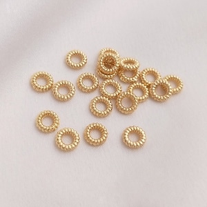 May include: A group of 20 gold jump rings, each with a textured, coiled design. The rings are scattered on a white surface.