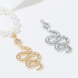 Op de afbeelding: Een gouden en zilveren slangenketting met een parelketting. De slangenhanger is versierd met heldere strasssteentjes.