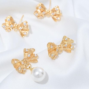 Puede incluir: Pendientes dorados con forma de lazo y detalles brillantes. Un pendiente presenta una perla blanca colgante. Los pendientes están colocados sobre un fondo blanco y sedoso, creando una estética lujosa.