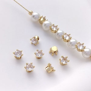 May include: A close-up of gold-toned jewelry findings with clear rhinestones. The findings are arranged on a white surface. There is a strand of pearls and gold beads in the background.