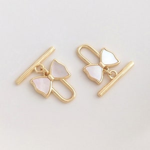 Könnte beinhalten: Zwei goldfarbene Toggle-Verschlüsse mit einem weißen Schleifen-Anhänger. Die Verschlüsse sind so konzipiert, dass sie als Verschluss für ein Kleidungsstück oder ein Accessoire verwendet werden können.