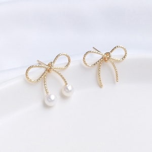 Pode incluir: Um par de brincos de laço de ouro com detalhes de pérolas. Os laços são feitos de fio trançado e as pérolas são brancas.