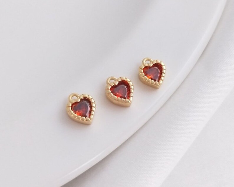 Puede incluir: Tres abalorios dorados en forma de coraz&oacute;n con detalles de gemas rojas. Los abalorios tienen un borde de cuentas y un bucle para sujetarlos a joyas.