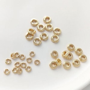 Può includere: Una collezione di piccoli elementi di gioielleria a forma di anello color oro. I componenti sono sparsi su una superficie bianca. Gli anelli hanno un aspetto strutturato e sono probabilmente utilizzati per la creazione di gioielli o l'artigianato.