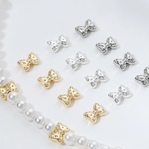 May include: A collection of silver and gold bow-shaped charms with a dotted texture. The charms are arranged on a white surface.