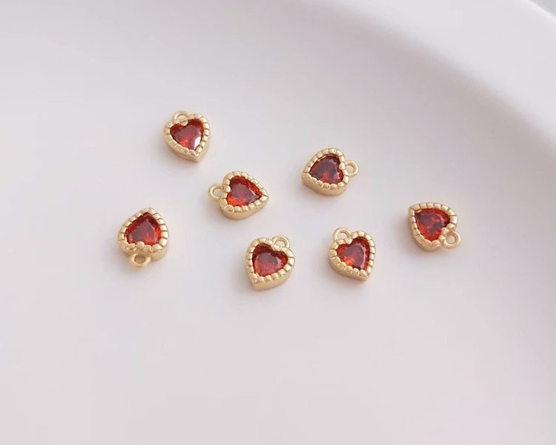 Puede incluir: Siete abalorios dorados en forma de coraz&oacute;n con centros de pedrer&iacute;a roja. Los abalorios tienen un bucle en la parte superior para sujetarlos a joyas.