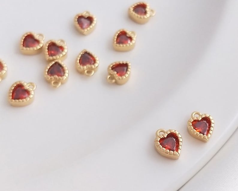 Puede incluir: Una colecci&oacute;n de peque&ntilde;os abalorios de coraz&oacute;n de oro con acentos de gemas rojas. Los abalorios tienen un bucle en la parte superior para sujetarlos a joyas u otras manualidades.
