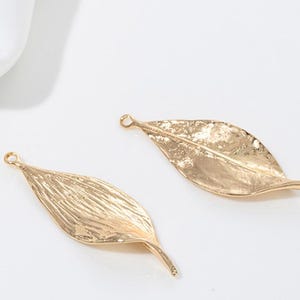 Op de afbeelding: Twee gouden blaadjes met een getextureerd oppervlak. De bedels hebben een lus aan de bovenkant om aan sieraden te bevestigen.