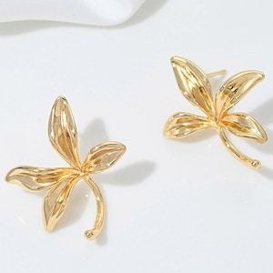 Può includere: Un paio di orecchini a bottone color oro. Ogni orecchino presenta un design a foglia stilizzato con tre petali curvi e un delicato stelo. Gli orecchini sono su uno sfondo bianco.