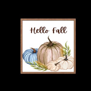 Op de afbeelding: Een ingelijst afdruk met de tekst "Hello Fall" in bruine letters. De afdruk toont een aquarel-illustratie van drie pompoenen: een blauwe, een beige en een witte. De pompoenen worden omringd door groene bladeren en tarwehalmen.