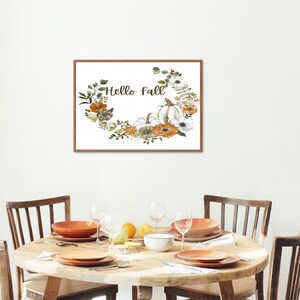 May include: A framed print with a white background and a floral wreath with pumpkins and the text "Hello Fall". The print is hanging on a white wall above a wooden table with four chairs.