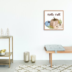 Op de afbeelding: Een ingelijst aquarel schilderij van pompoenen en pompoenen met de tekst "Hello Fall" in bruine letters. De pompoenen zijn wit, beige en blauw.